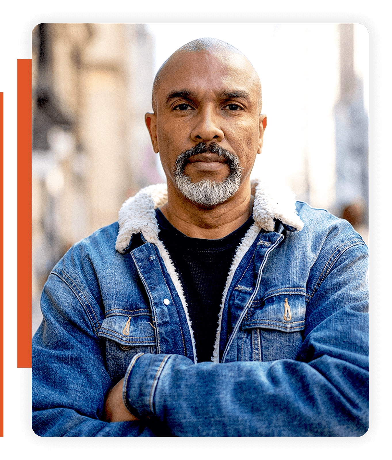 A man with a salt-and-pepper beard wearing a denim jacket with a shearling collar, standing with arms crossed on a city street