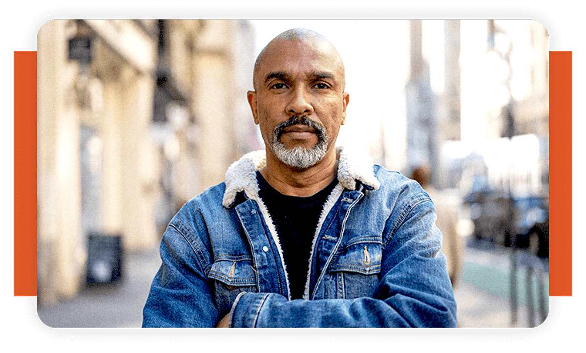 A man with a salt-and-pepper beard wearing a denim jacket with a shearling collar, standing with arms crossed on a city street.