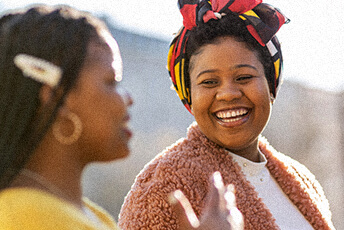Two women smiling and conversing outdoors, with one wearing a colorful headscarf and the other wearing a yellow sweater.