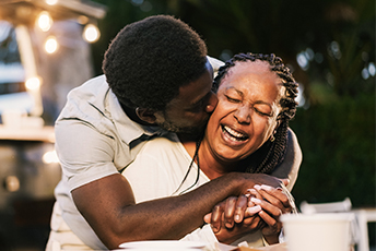 A man hugging a woman from behind and kissing her cheek, both laughing joyfully