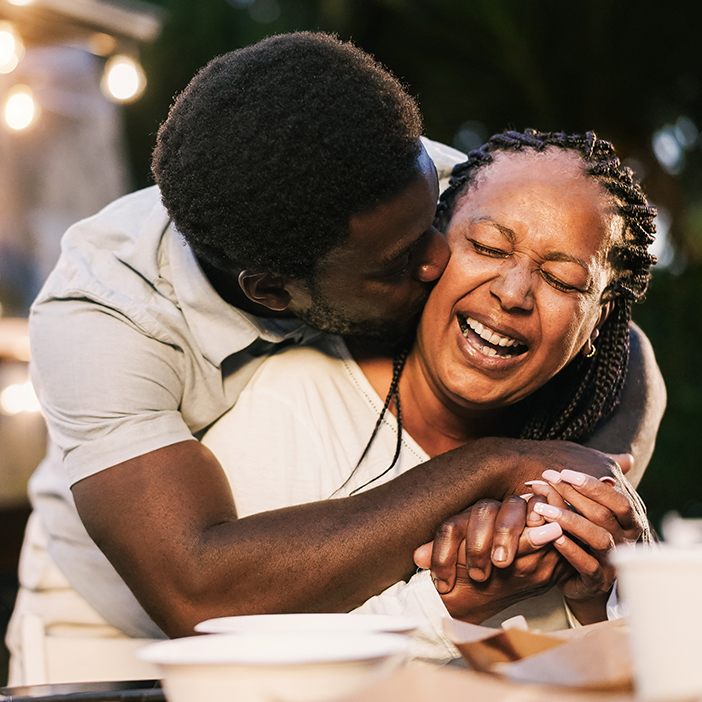 A man hugging a woman from behind and kissing her cheek, both laughing joyfully