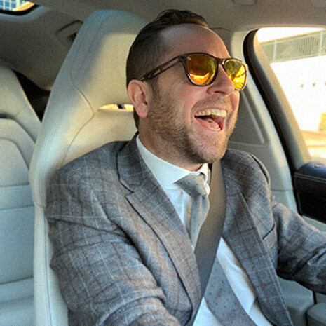 A man in a suit and sunglasses laughing while driving a car