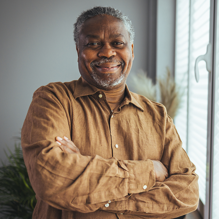 A man with gray hair and a beard, wearing a brown shirt and smiling with his arms crossed