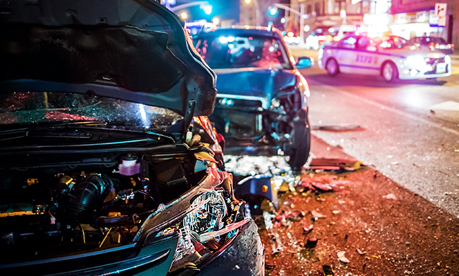 Two heavily damaged cars on a city street at night with police cars in the background