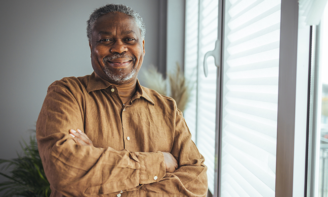 A man with gray hair and a beard, wearing a brown shirt and smiling with his arms crossed